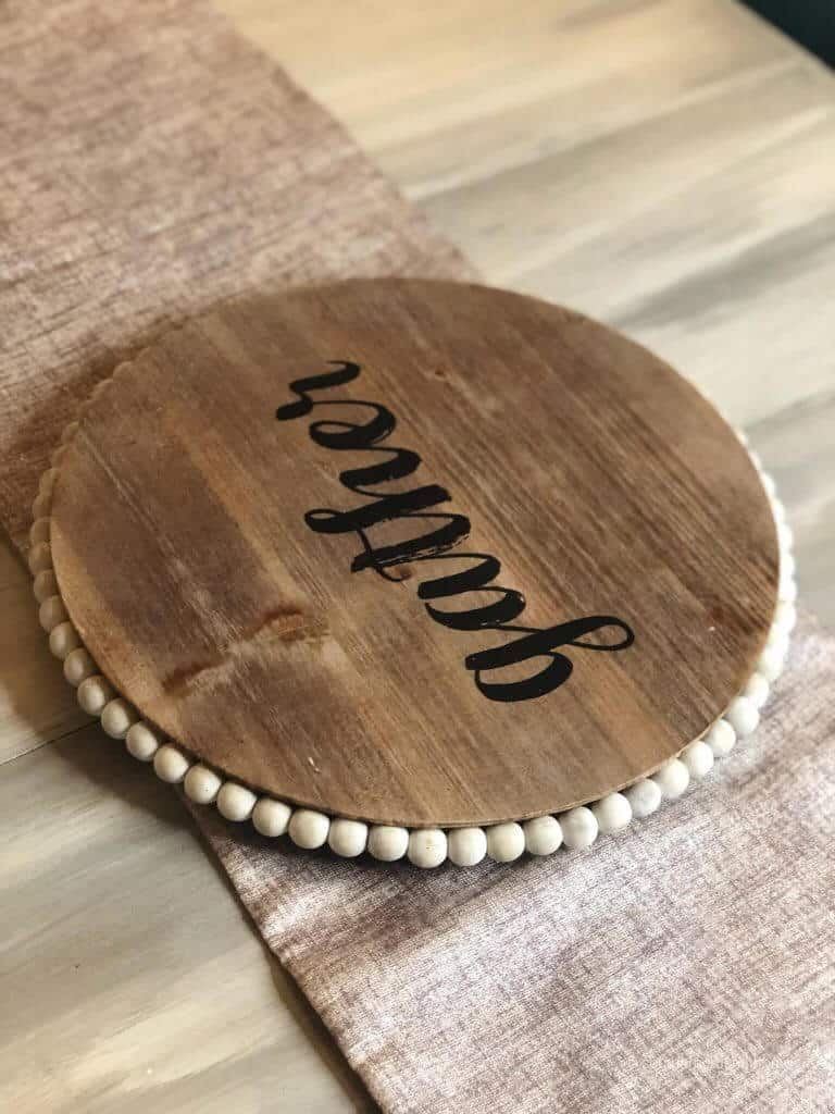 A round piece of wood with the word gather on it and wooden bead trim