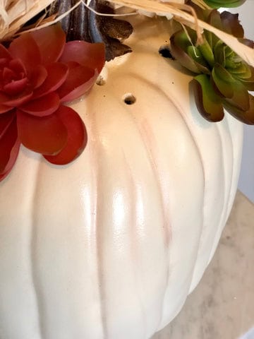 A white faux pumpkin with a brown spot and holes where faux succulents are placed.