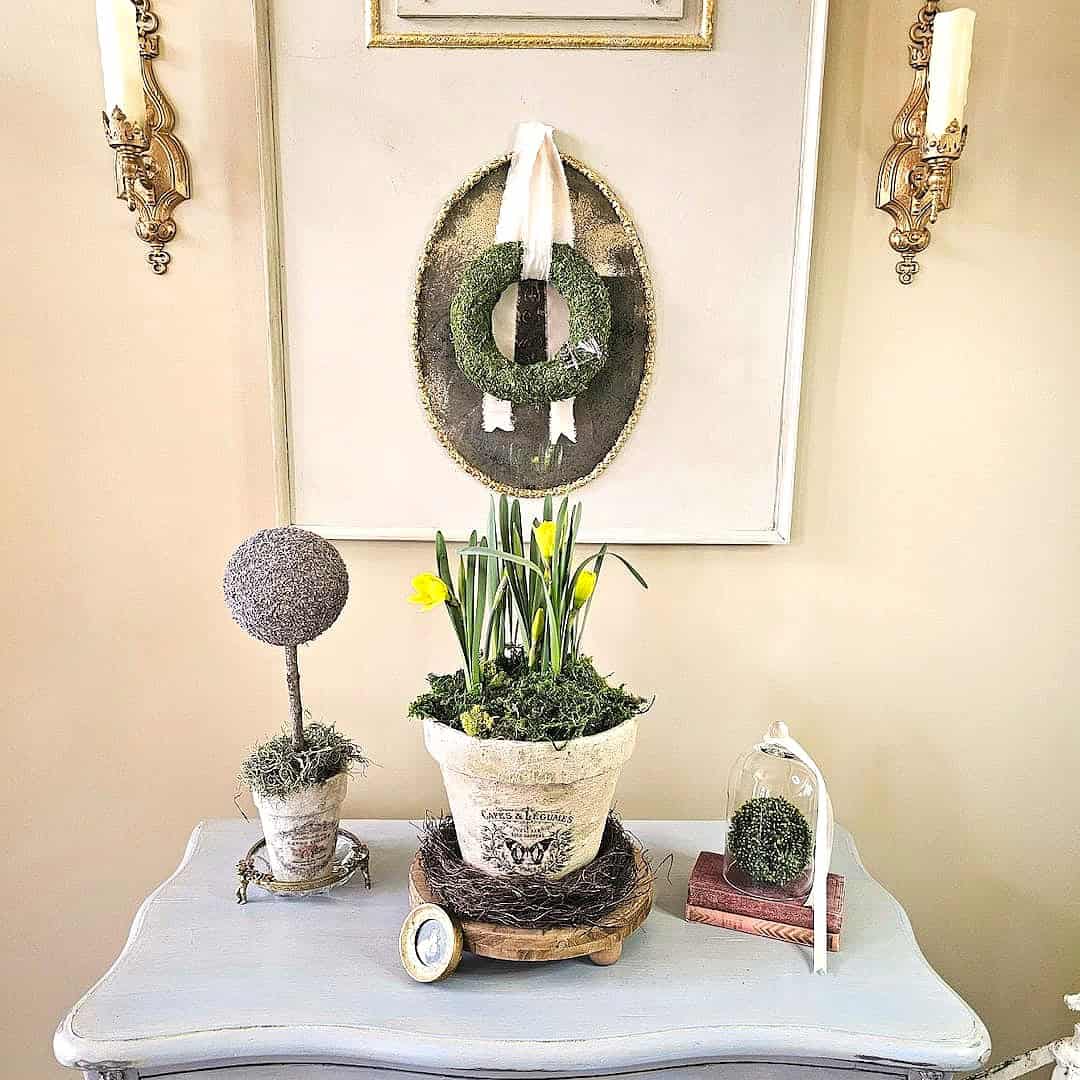 A light blue table holds a potted plant with yellow flowers, a moss topiary, a small clock, and a glass cloche covering a moss ball; above hangs a green wreath on a white-framed backdrop&mdash;perfect for any Home Tour Code inspiration.