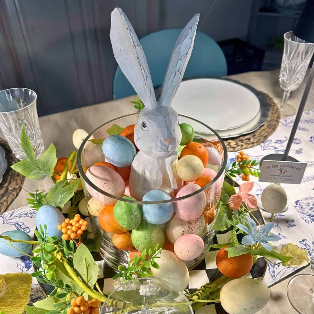 A white ceramic bunny sits in a glass bowl filled with pastel eggs, surrounded by artificial flowers and greenery on a decorated table set for a meal&mdash;perfect for your next Home Tour celebration.