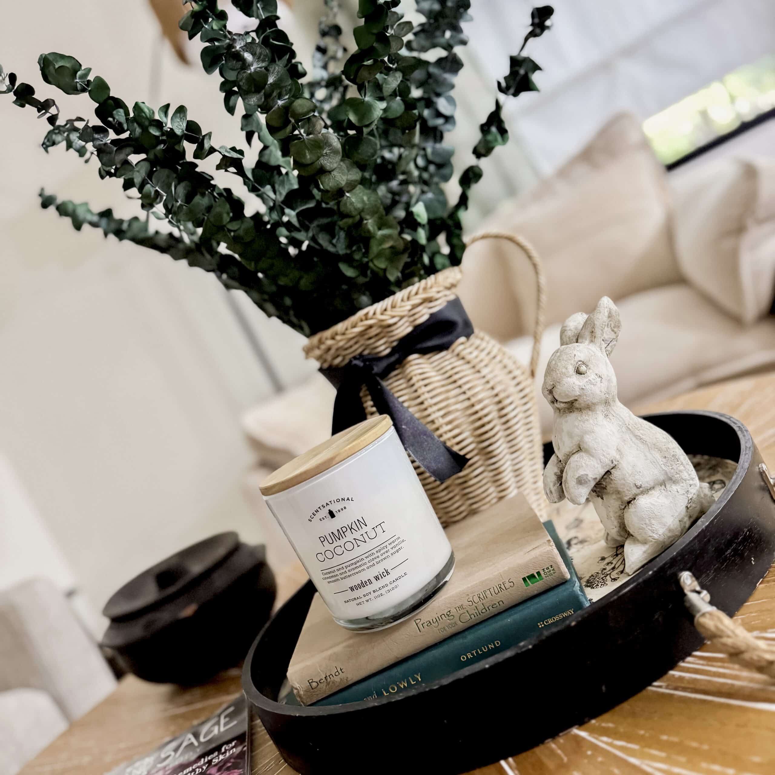 A decorative tray on a wooden table holds a white candle, two stacked books, a ceramic rabbit figurine, and a woven vase with green foliage&mdash;perfect for any Home Tour in a cozy, light-filled living room.