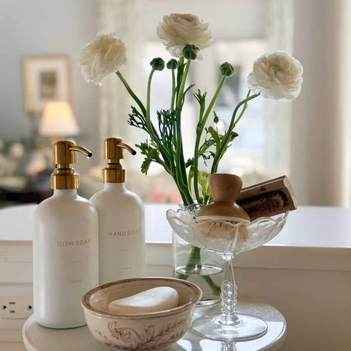 A neatly arranged countertop with bottles of dish soap and hand soap, a bowl with a bar of soap, a glass dish holding brushes, and a vase of white ranunculus flowers&mdash;this space is pure Home Tour Code inspiration.