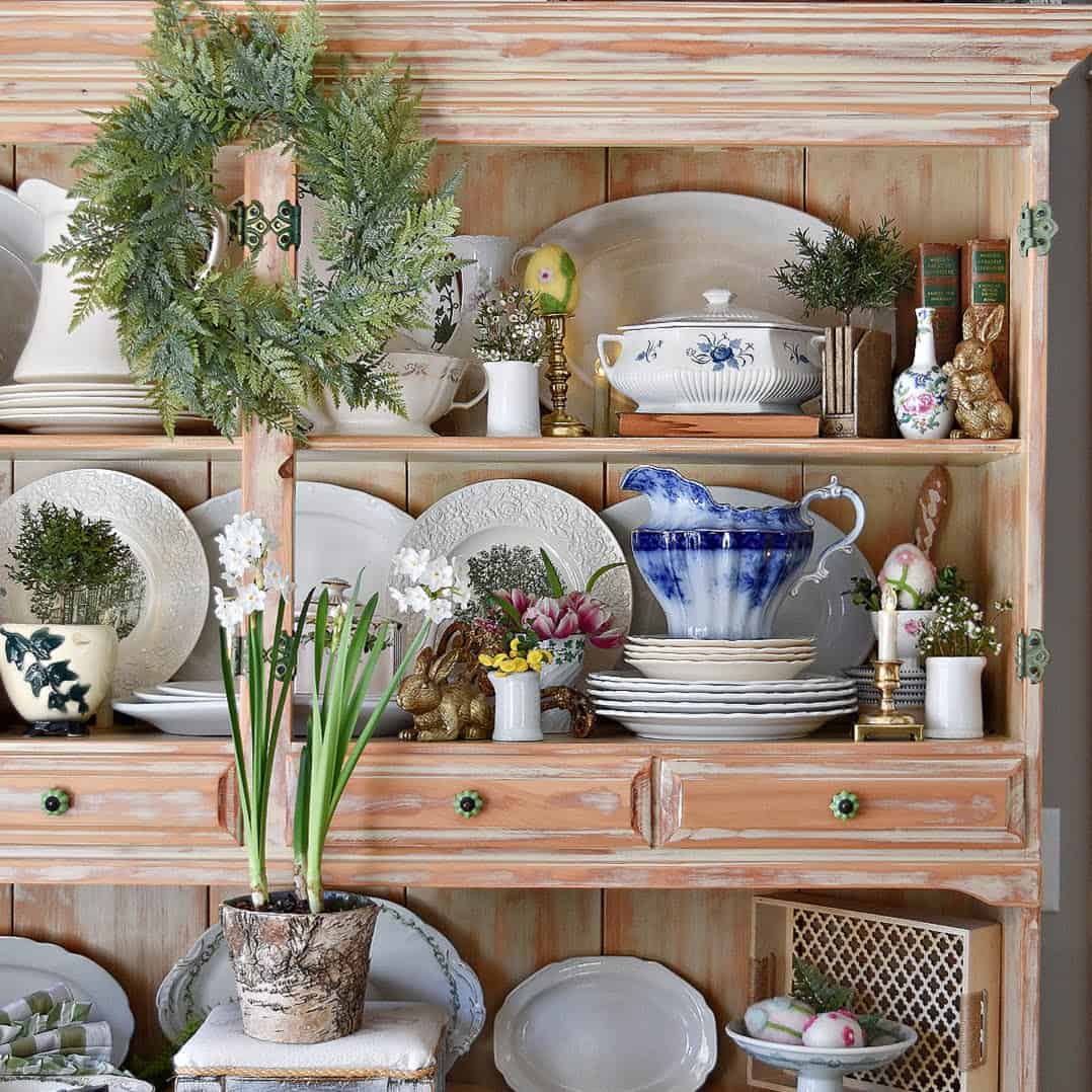 A rustic wooden hutch displays white and blue china, plates, teapots, and cups, decorated with a green wreath, potted flowers, bunny figurines, and Easter eggs&mdash;perfect for a spring-themed Home Tour arrangement.