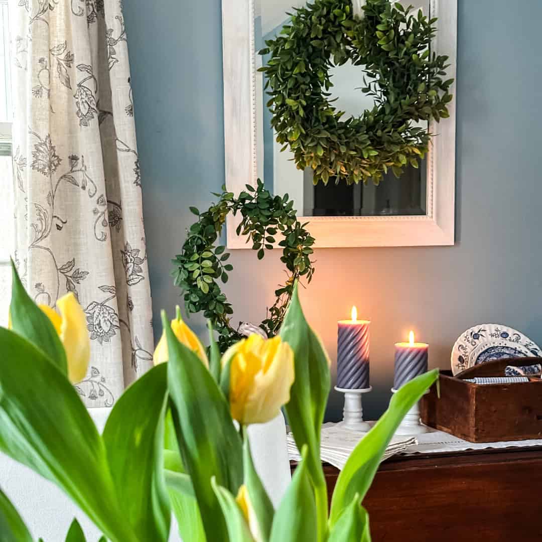 A cozy room with yellow tulips in the foreground, two green wreaths&mdash;one hanging on a mirror and one leaning on the wall&mdash;and two lit candles on a table beside a wooden box and blue-patterned plates, ready for a Home Tour.