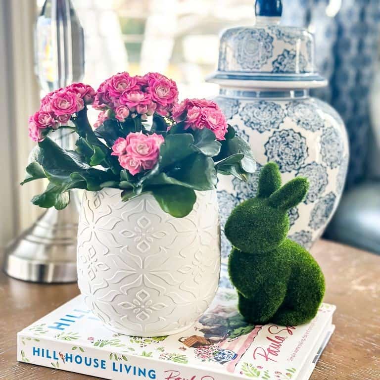 A white textured vase with pink flowers sits on two stacked books next to a green moss bunny and a blue-and-white patterned jar, styled for the perfect Home Tour Code look on a tabletop with soft natural light.