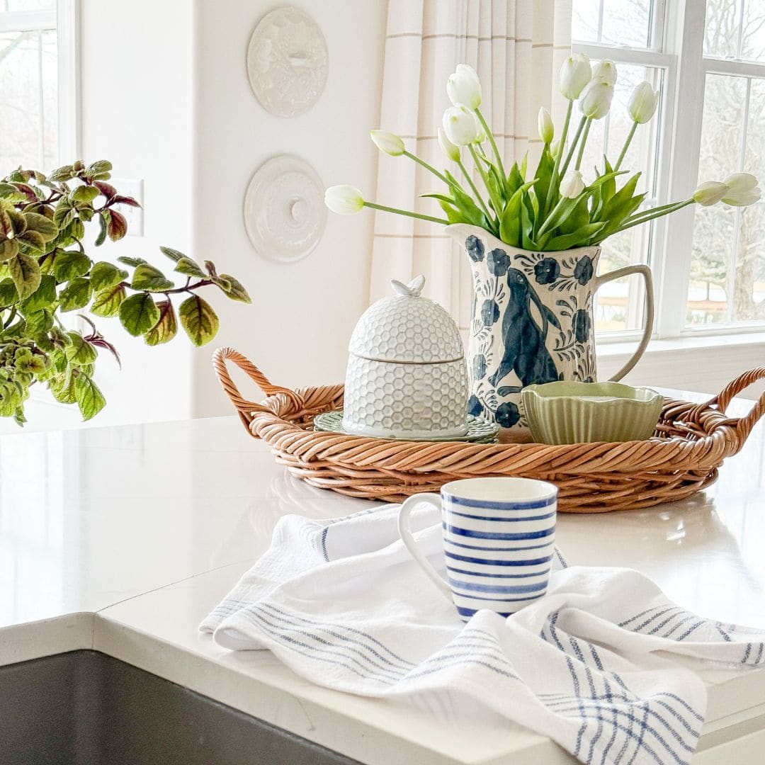 A wicker tray with a floral pitcher of white tulips, a honey jar, and green bowls sits on a white countertop. In front, a striped mug and blue-striped towel are by the sink. Natural light fills the space&mdash;perfect for your next Virtual Home Tour.
