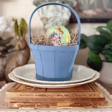 A small blue basket with an Easter egg decorated with a bunny and flowers sits on a white plate, atop vintage magazines&mdash;an inviting Home Tour scene, with a plant and brown bunny figurine visible in the blurred background.