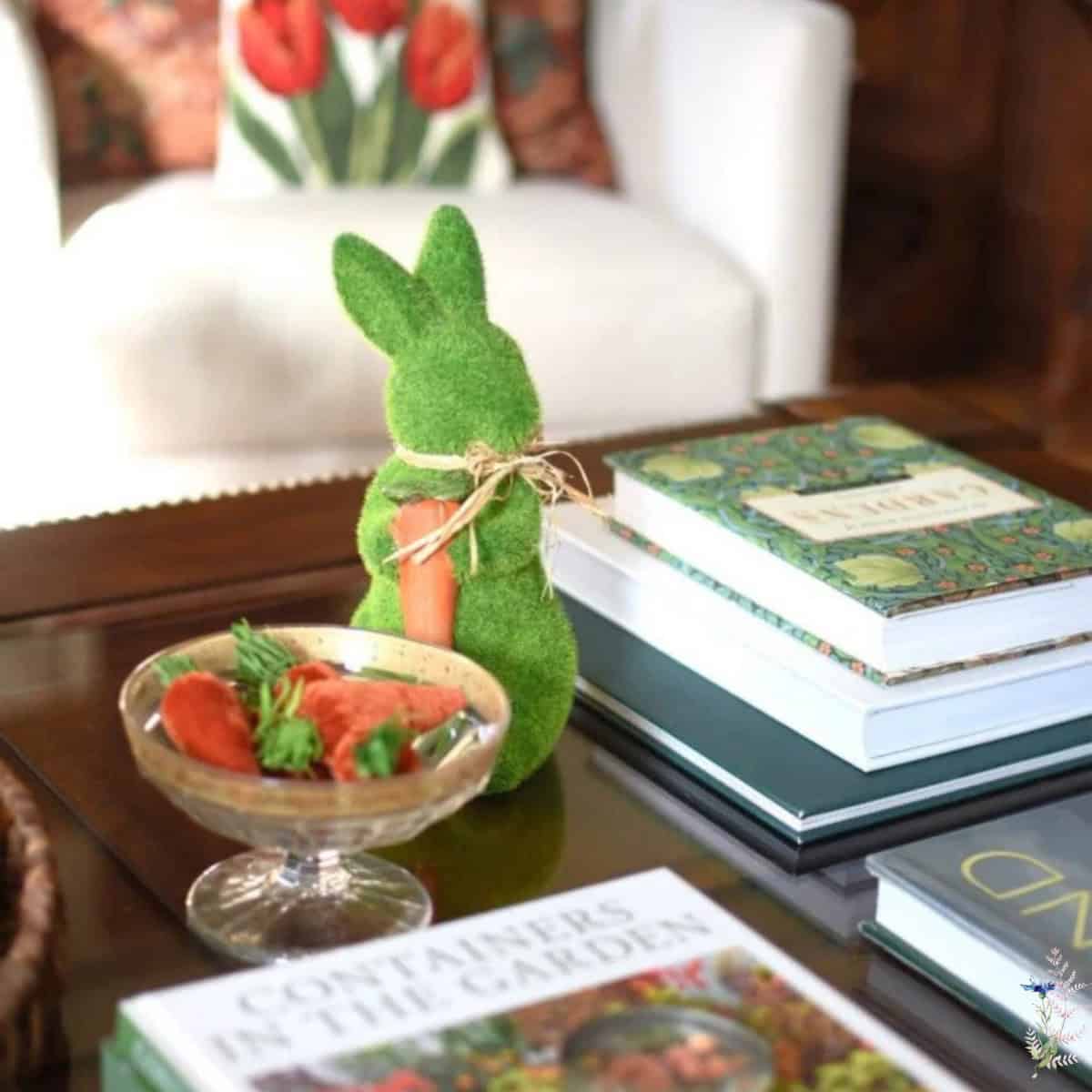 A green, grass-like bunny figurine with a twine bow and carrot sits on a coffee table near books and strawberries&mdash;perfect for styling your space or sharing in your next virtual home tour. A chair with a tulip pillow accents the background.